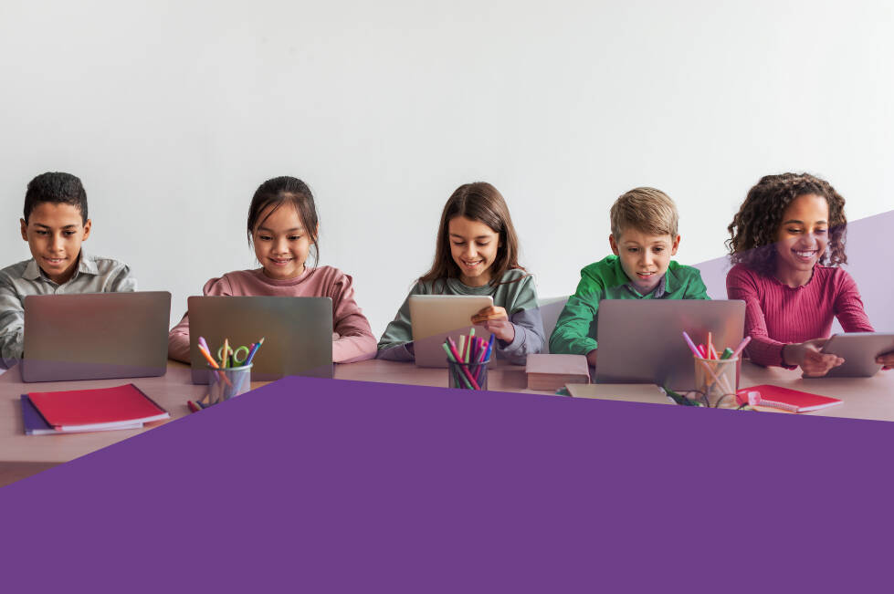 Five elementary-aged students sit side by side at a table using laptops and tablets. The children are smiling and engaged with their devices, with school supplies such as notebooks and cups of colored pencils on the table.